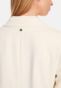 Cream-colored coat with a folded collar, featuring a metal accent button at the back, smooth texture, and structured design.