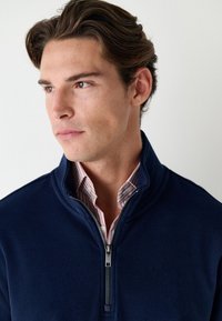 Námořnický fleecový svetr na zip s vysokým límcem a stříbrným zipem. Nositelný přes světle růžovou a bílou pruhovanou košili, vykreslující vrstvený vzhled.