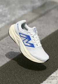 Chaussure de sport blanche avec des accents bleus et un dessus texturé, dotée d'une semelle épaisse, d'un système de laçage et d'un logo visible.