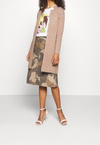 Vrouw in een beige lange cardigan, witte graphic t-shirt, knopenlijf camouflage rok en witte hoge hakken, staand voor een effen achtergrond.