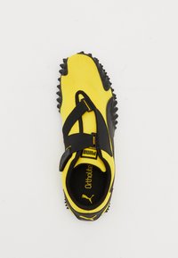 Yellow athletic shoe with black accents, featuring a textured fabric upper, adjustable strap, and rubber outsole with protruding traction studs.