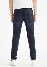 Sötét farmer skinny nadrág elülső derékrésszel, két hátsó zsebbel és sima textúrával, fehér tornacipővel párosítva.