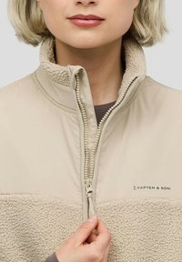 Beiger Fleece-Jacke mit einem strukturierten unteren Teil, glattem oberen Teil und Reißverschluss. Mit Kapten & Son-Logo auf der Brust.