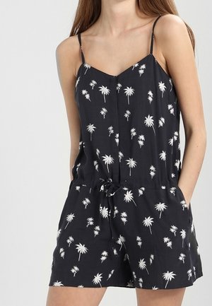 Vrouw draagt een zwarte mouwloze romper met witte palmboomprints en een strik in de taille, handen in de zakken.