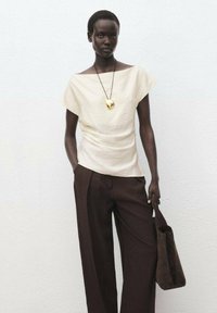 Haut crème, à manches courtes et coupe ample, associé à un pantalon large marron foncé et un sac fourre-tout assorti. Collier en pendentif doré en accent.