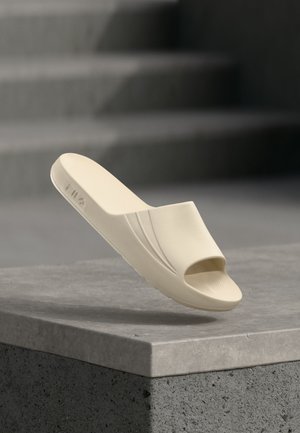 Bézs slip-on szandál nyitott orral, texturázott talppal és FILA logóval az oldalon, betonperem felett, háttérben lépcsőkkel.