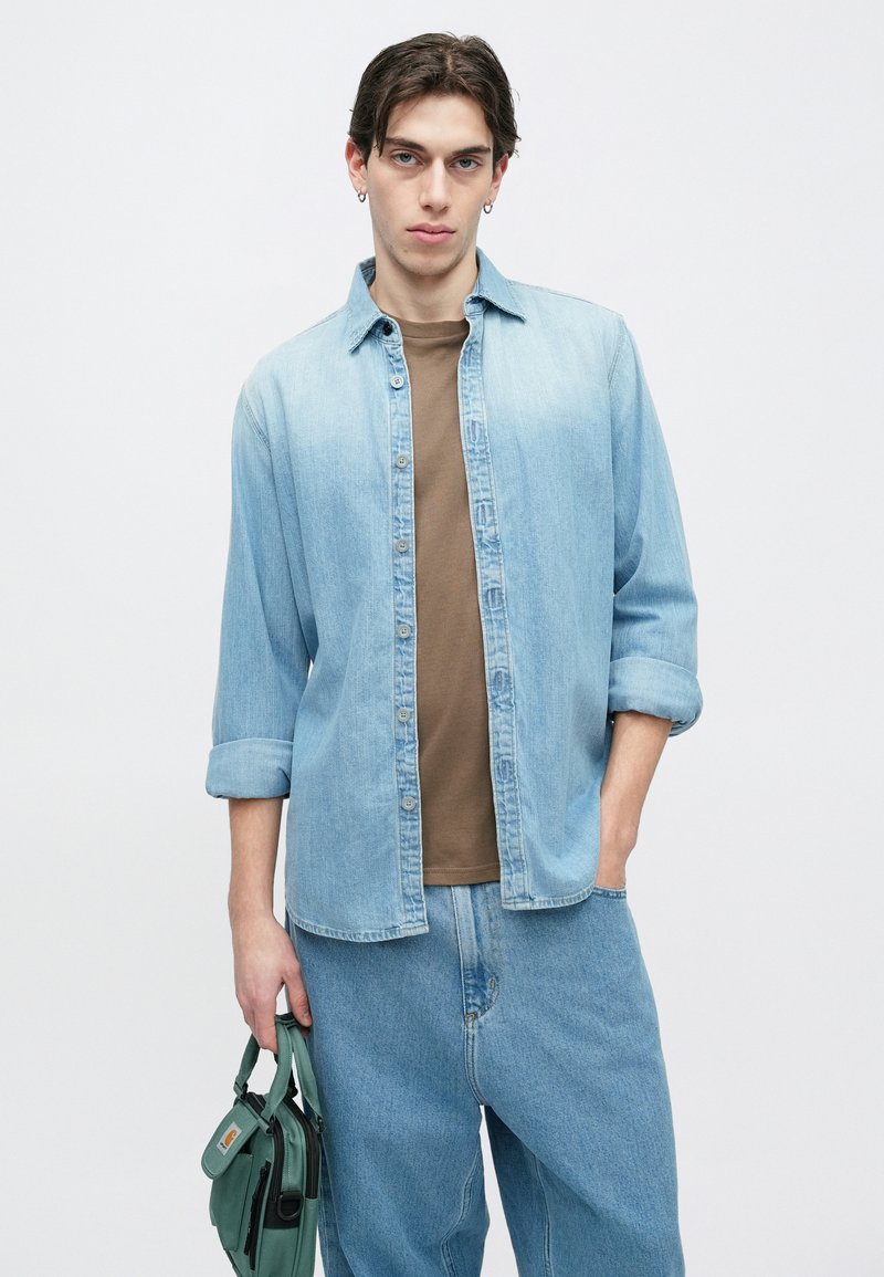 Camisa vaquera azul claro, de corte holgado, abotonada con las mangas remangadas. Camiseta marrón debajo, combinada con vaqueros azules sueltos. Bolso de hombro verde.