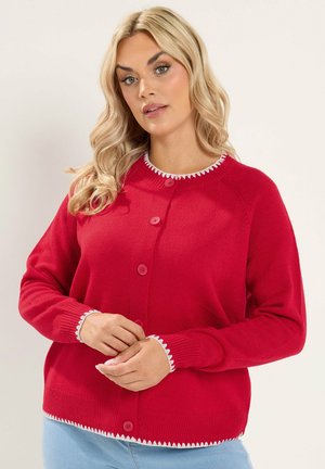 Woman with blonde hair wearing a red long-sleeve cardigan with white trim and blue jeans, standing against a neutral background.