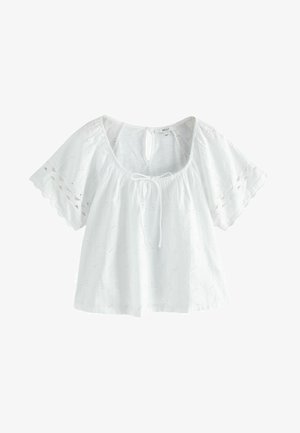 Blouse en coton blanc avec des manches courtes évasées, ornée de broderies en œillet et d'un dos en forme de trou de serrure, avec un col noué et un tissu texturé.