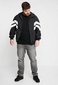 Sort zip-up jakke med hvide chevronstriber, stylet med en sort T-shirt og grå jeans. Jakken har hætte og elastiske manchetter.