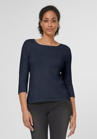 Dunkelblaues, gestricktes Langarmoberteil mit weitem Rundhalsausschnitt und tailliertem Design. Glatte Textur und dezenter Stretch, kombiniert mit dunklen Jeans.