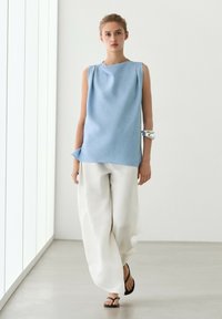 Giovane donna che cammina in casa indossando un top azzurro senza maniche, pantaloni bianchi larghi, sandali neri e un braccialetto d'argento al braccio sinistro.