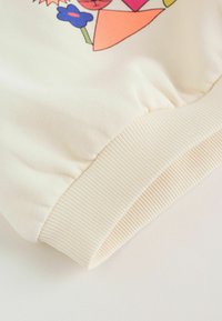 Tela de color crema claro con dobladillo acanalado. Presenta patrones geométricos de flores coloridas. Textura suave, diseño casual, manga con puño elástico fruncido.