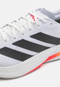 Chaussure de sport avec un dessus en maille blanche, des rayures texturées noires et une semelle extérieure en dégradé de couleurs. Dispose de trous de lacets renforcés et d'un col rembourré.