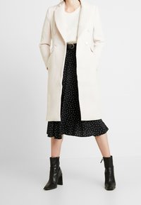 Abrigo beige de doble botonadura con solapas de muesca, acompañado de una falda negra con lunares y botines negros de tacón. Tejido suave, diseño de longitud media.
