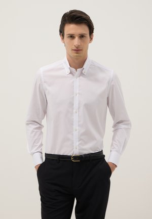 Uomo in piedi con le mani nelle tasche, indossa una camicia bianca con bottoni e pantaloni neri con cintura nera su uno sfondo semplice.