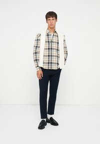 Geruit overhemd met een knoopsluiting in beige, marineblauw en oranje tinten, gecombineerd met donkere broeken en zwarte instappers, aangevuld met een crèmekleurige trui over de schouders.