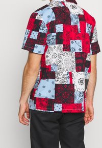 Camicia a maniche corte con un design a patchwork che presenta motivi paisley rossi, neri, azzurri e bianchi, realizzata in tessuto leggero.