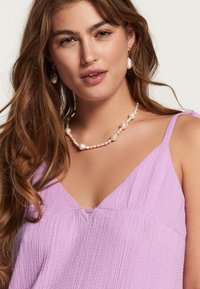Top fără mâneci de culoare violet deschis, cu un material texturat, având un decolteu în V și un detaliu mic în formă de cheie. Purta un colier cu perle.