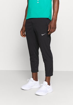 Mężczyzna ubrany w czarne spodnie sportowe Nike, białe buty sportowe i turkusową koszulkę, stoi na jasnej podłodze na tle gładkiej ściany.
