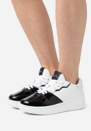 Zapatillas en blanco y negro con parte superior de charol, acentos texturizados, cordones blancos, suela gruesa y una etiqueta de logo oscura en la lengüeta.