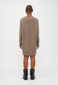 Vestido de malha castanho com um corte solto, mangas longas e punhos canelados, combinado com botas de tornozelo pretas com tiras decorativas.