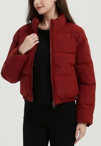 Doudoune rouge avec un col montant, fermeture éclair à l'avant et taille élastique. Présente un rembourrage cousu et des poches latérales. Associée à un haut noir.