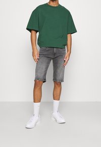 Camiseta verde de gran tamaño, pantalones cortos de mezclilla grises, zapatillas blancas y calcetines blancos. Tiene mangas cortas y un ajuste casual.