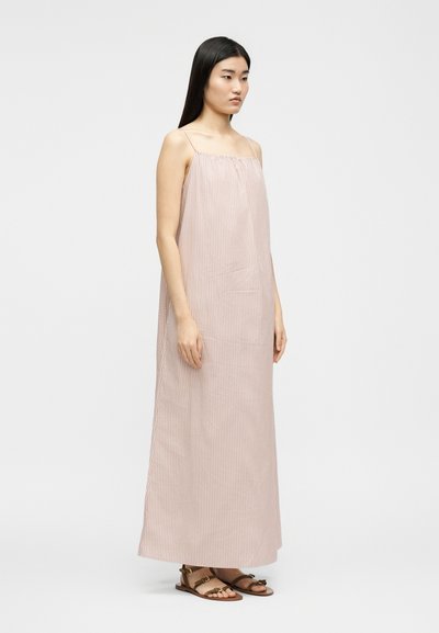 Γυναίκα που στέκεται φορώντας ένα μακρύ, χωρίς μανίκια, ροζ και λευκό ριγέ φόρεμα με λεπτές τιράντες και καφέ επίπεδες σανδάλια.