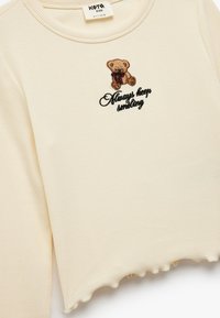 Camiseta de manga larga de color crema con ribetes y dobladillo con puntilla, que presenta un oso de peluche bordado y el texto "Siempre sigue sonriendo."