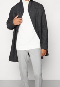 Czarny płaszcz w warkocz z szalowym kołnierzem, z czarnymi guzikami, nałożony na biały sweter, w zestawie z jasnoszarymi spodniami.