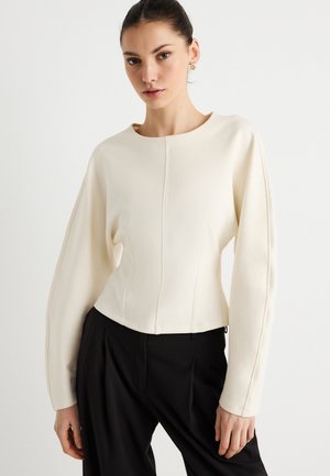 Fehér, rövidített hosszú ujjú felső, sima anyagból, kerek nyakkivágással és varrási részletekkel, fekete, pliszírozott nadrággal párosítva.