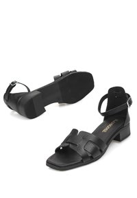Sandalias de cuero negro con acabado texturizado, punta abierta y correa al tobillo. Presentan un tacón cuadrado y dos tiras superpuestas en la parte delantera.