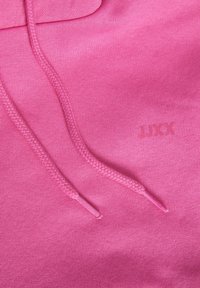 Gros plan sur le tissu rose d'un sweat à capuche montrant des cordons tressés et un logo JXX subtilement embossé au niveau de la poitrine.