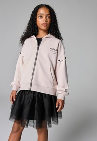 Ružičasti hoodie na zip s velikim prednjim džepovima i crnim detaljima, u kombinaciji s crnom til suknjom sa slojevitim dizajnom.