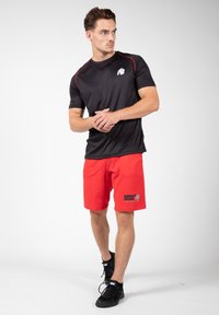 Zwart sportshirt met rode accenten; rode shorts met een logo. Draagt zwarte schoenen en staat in een neutrale pose.