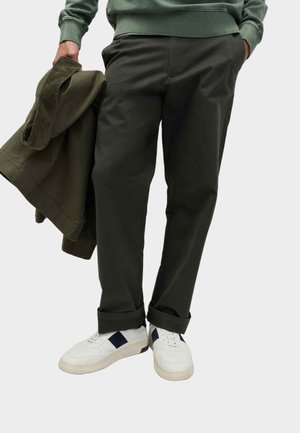 Person iført mørkegrønne bukser og hvide sneakers, der holder en matchende mørkegrøn jakke i den ene hånd, står mod en ensfarvet baggrund.