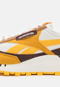 Reebok-sneaker med ett vitt textilt ovanmaterial och gult och brunt mockaaccent, runda snören och en texturerad gummisula.