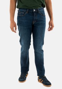 Jeans in denim blu scuro con vestibilità a gamba dritta, design a cinque tasche e dettagli di cucitura sottili. Abbinati con sneakers nere e una camicia verde.