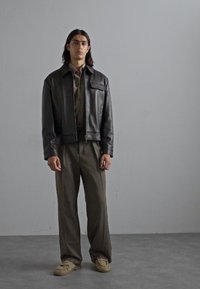 Black leather jacket with a front pocket, paired with olive green shirt and matching trousers. Beige shoes complete the outfit.