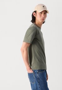 T-shirt en coton vert olive, col rond, manches courtes, associé à un jean en denim bleu. Casquette beige avec un emblème sur le devant. Fond uni.