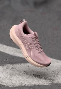 Light pink running shoe with laces, cushioned sole, and white logo, captured mid-air above a paved surface with white markings.