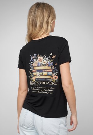 Frau mit langen blonden Haaren, die ein schwarzes T-Shirt trägt mit gestapelten Büchern, Blumen und Schmetterlingen sowie dem Schriftzug "BOOKTROVERT" auf dem Rücken.