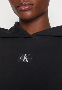 Czarna prążkowana bluza z kapturem z troczkami. Z przodu naszywka w kształcie kwadratu z logo Calvin Klein Jeans w bieli i szarości.