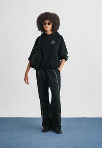 Νεαρός με σγουρά μαλλιά, που φοράει γυαλιά ηλίου, μαύρη φούτερ μπλούζα με κουκούλα, μαύρο αθλητικό παντελόνι με λευκές ρίγες, μαύρα παπούτσια και κρατά μια μαύρη τσάντα ώμου.