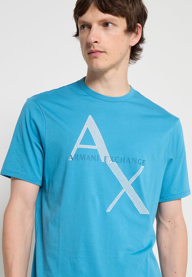 Giovane uomo che indossa una maglietta Armani Exchange di un brillante blu con grandi lettere bianche "AX" e un testo più piccolo "Armani Exchange" sul petto.