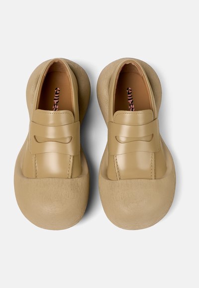 Zapatos slip-on beige con punta redondeada y suela exterior de goma texturizada. La parte superior de cuero suave presenta una correa decorativa y detalles de costura.