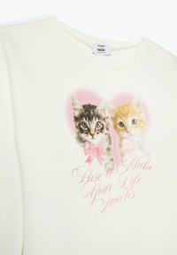 Crème sweatshirt met twee katten met roze strikken, omringd door een hartvorm. De tekst luidt: "Hier om je leven zoeter te maken."