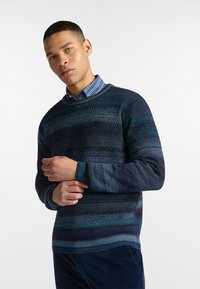 Maglione lavorato a maglia di colore blu scuro con strisce orizzontali in diverse tonalità di blu e verde acqua; scollo tondo, texture morbida e maniche lunghe.