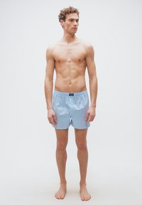 Νεαρός άνδρας που στέκεται ξυπόλυτος, φοράει ανοιχτό μπλε και λευκό ριγέ μποξέρ και κοιτάζει ελαφρώς προς τα δεξιά με φόντο ένα απλό υπόβαθρο.